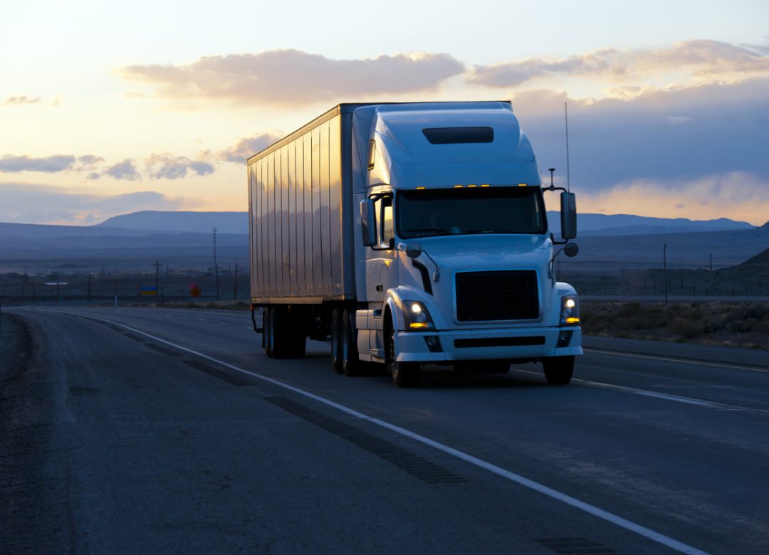 Huge modern big big semi truck with a trailer moving on the road in the evening twilight with headlights on the background of the picturesque hills with reflection burning rays of the evening sunset.